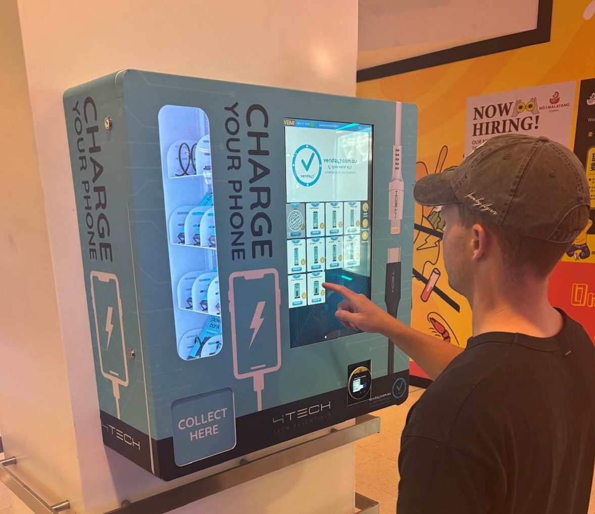 A wall-mounted teal 4Tech Charge Your Phone machine at a live venue, a customer choosing a product on the touchscreen with a charging cable extended for use